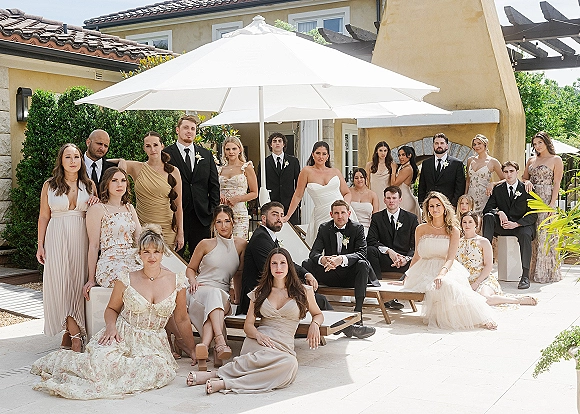 Wedding party portrait with bride and attendants in bridesmaid dresses and black suits, posed on patio lounge chairs by a villa pergola