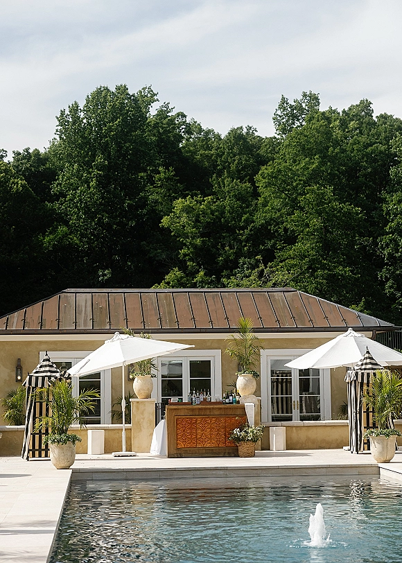 Outdoor wedding bar with liquor bottles and bar tools, styled as a wedding cocktail bar setup beside the pool under white umbrellas