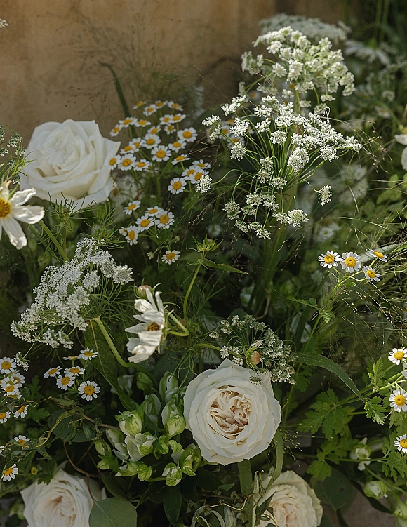 Wedding florals with wildflower wedding flowers, white roses and chamomile accented with airy greenery against a stone wall backdrop