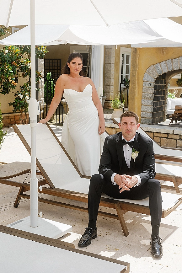 Couple portrait of bride and groom posing, bride standing by seated groom in tux, on a stone patio at a villa with arches
