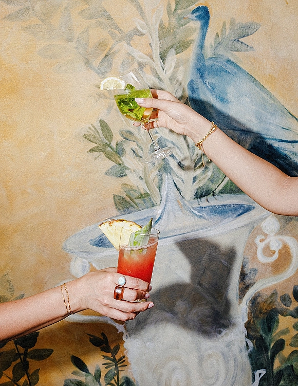 Wedding cocktails held up for a toast, two colorful signature wedding drinks with citrus garnish in front of a painted mural wall