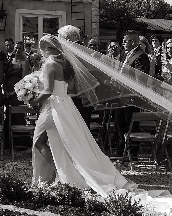 Wedding processional as the bride walking down aisle with long veil and white bouquet, groomsmen in suits and guests seated in courtyard chairs