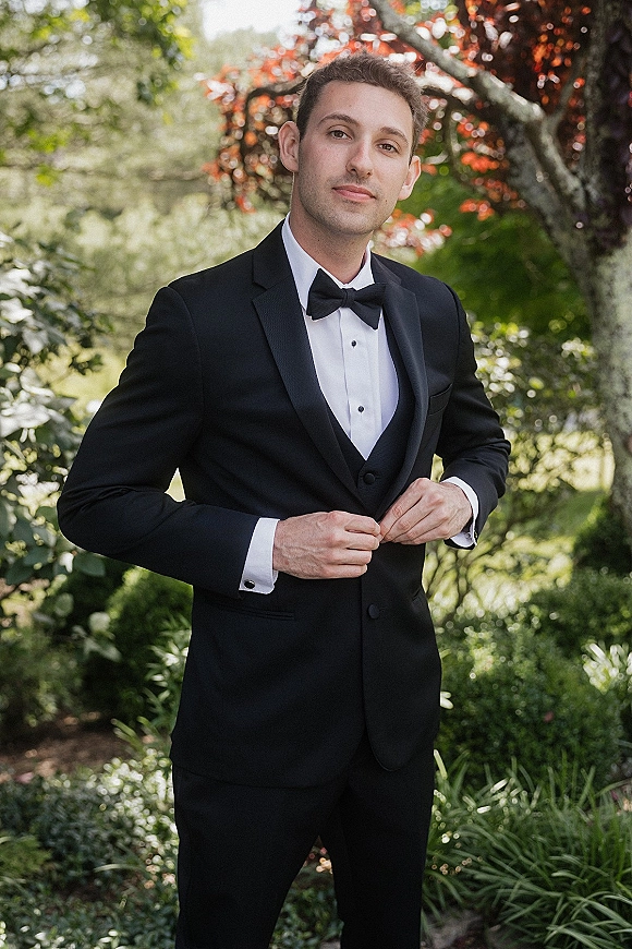 Groom portrait in a black tuxedo groom look with bow tie and waistcoat, standing in natural light amid lush garden foliage