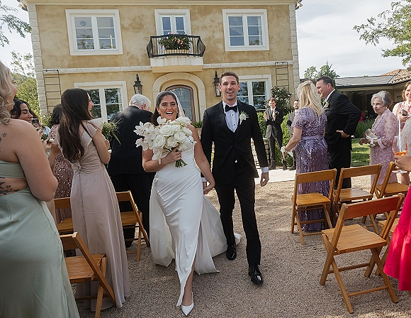 Wedding recessional as bride and groom walk hand in hand past guests, bride in strapless gown holding white rose orchid bouquet in villa courtyard aisle