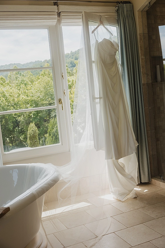 Wedding dress on hanger with bridal veil, softly lit by a large window in a tiled bathroom with bathtub and curtains