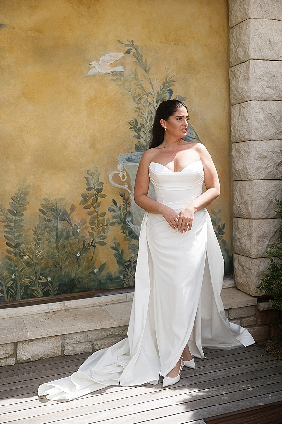 Bridal portrait of a bride in a strapless wedding dress with satin train and high ponytail, leaning by a painted mural wall and stone backdrop