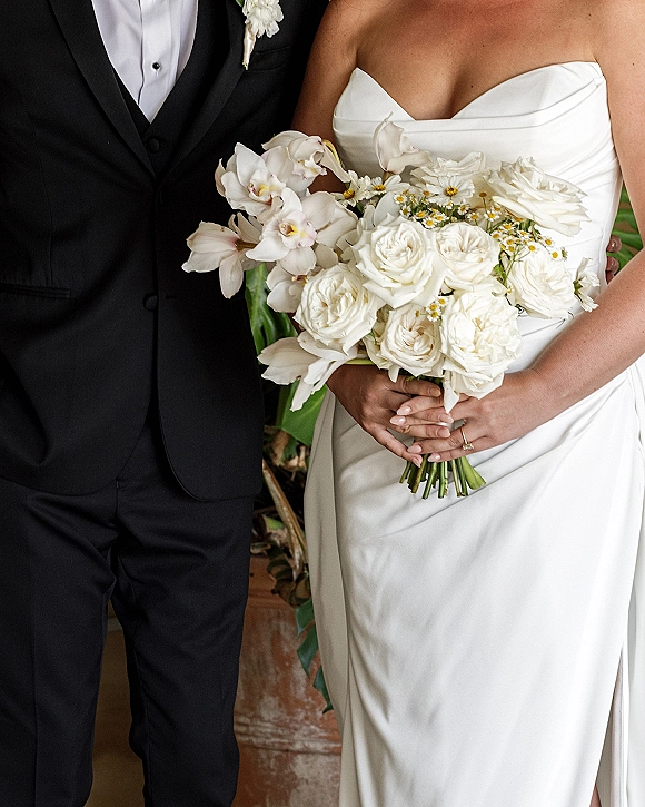Bride and groom portrait with a white rose bouquet and orchids, her strapless gown beside his black tuxedo, indoors by plants