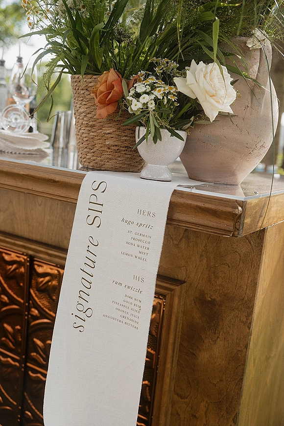Wedding bar menu fabric banner with calligraphy signature drinks sign on a wooden bar cart, framed by potted daisies and roses outdoors