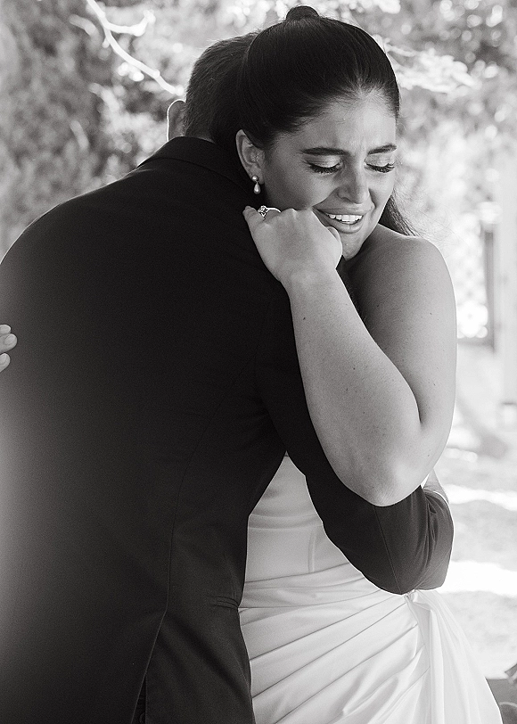 Wedding hug as bride hugging groom, eyes closed, showing engagement ring and pearl drop earrings in soft daylight by trees
