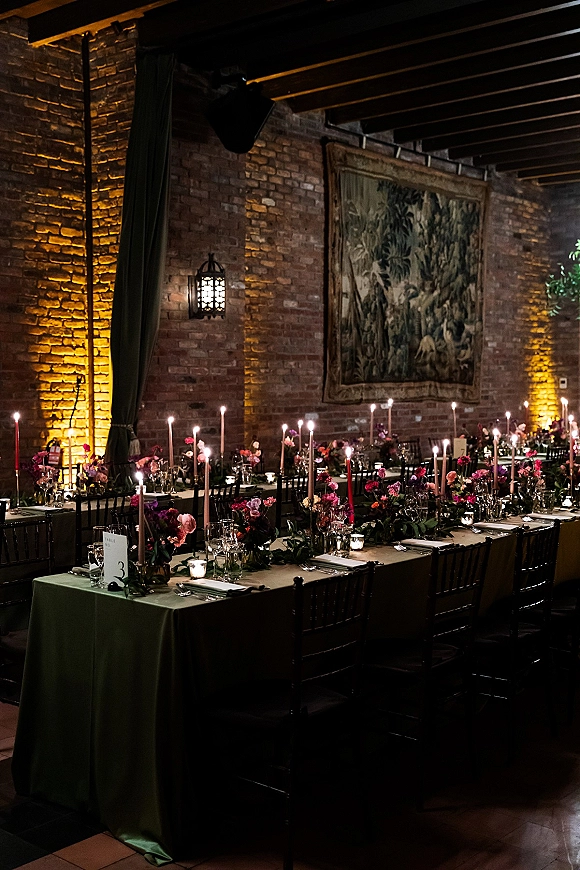 Reception tablescape with candlelit wedding reception glow, taper candles and dark floral centerpieces on a long table against exposed brick and tapestry