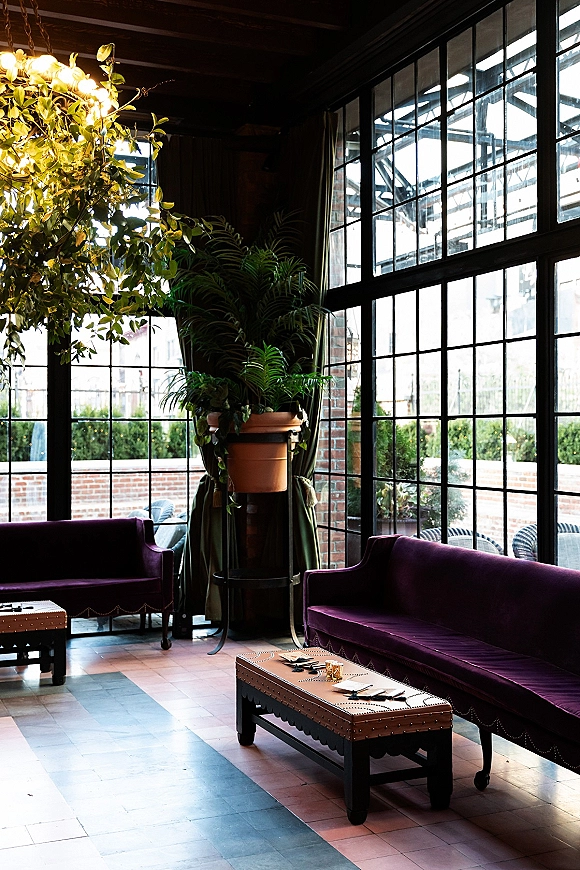 Wedding lounge seating with velvet sofas and coffee tables, lit by candles beneath a chandelier, set against industrial windows and brick wall backdrop