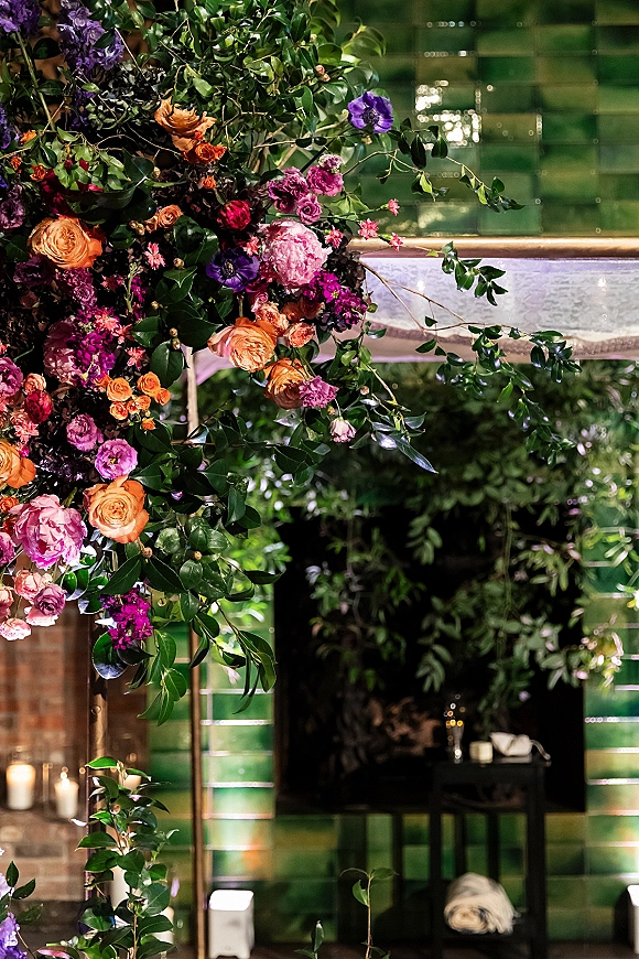 Wedding floral installation with hanging floral arrangement of roses and peonies on a gold frame, with candles before a green tiled fireplace wall
