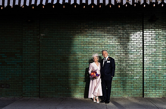 Couple portrait of an older wedding couple, senior bride and groom by a green brick wall, bride holding a colorful bouquet under string lights
