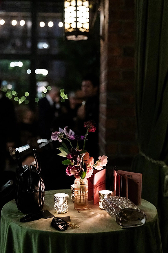 Cocktail table decor with ribbed glass vase flowers, votive candles, menus and table number beside a clutch in dim lantern-lit reception space