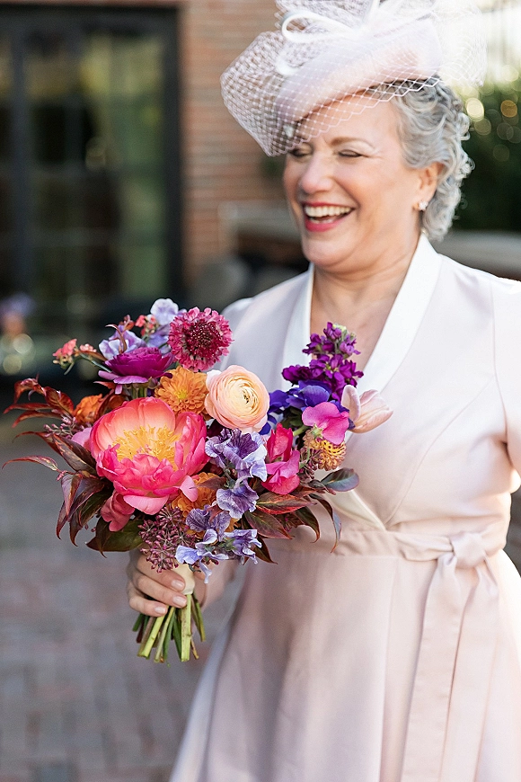 Bridal portrait of an older bride laughing with a colorful bouquet, wearing a birdcage veil fascinator on an outdoor brick patio with greenery