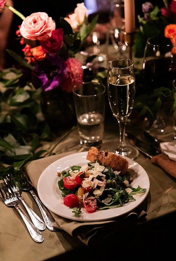 Reception tablescape with wedding place setting, pink-red floral centerpiece, taper candles, champagne flute, plated salad and bread in dim light
