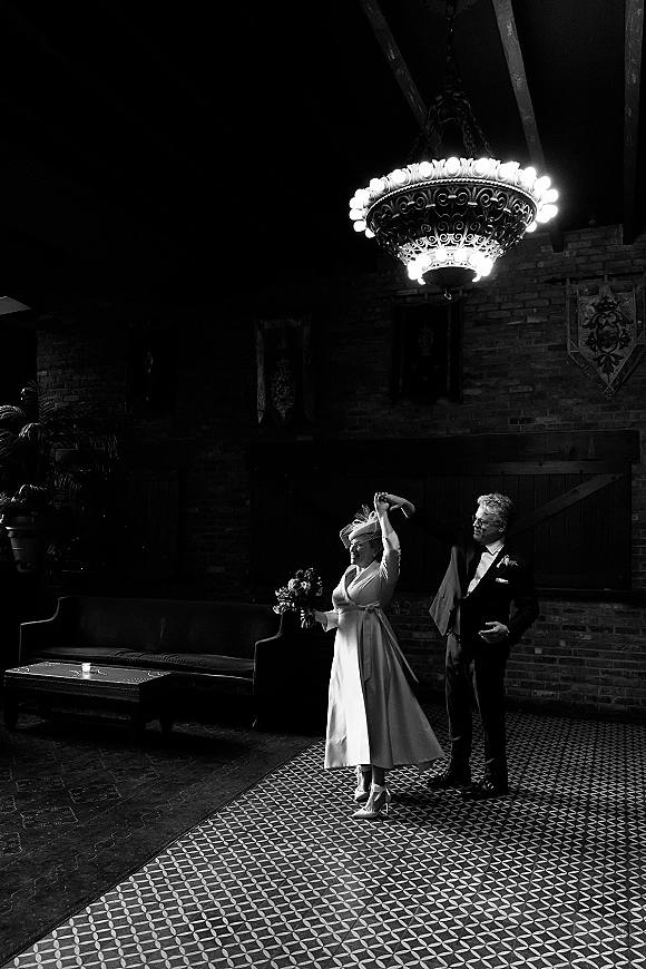 Wedding dance moment as bride twirls in a tea-length dress holding a bouquet, groom in tux watches under a chandelier by brick wall