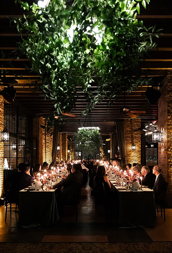 Reception tablescape with a long banquet table wedding set in candlelight, taper candles and floral centerpieces beneath hanging greenery in a brick venue