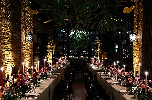 Reception tablescape on a long banquet table with taper candles, pink and purple florals, and greenery garland in a brick-walled, windowed space at night