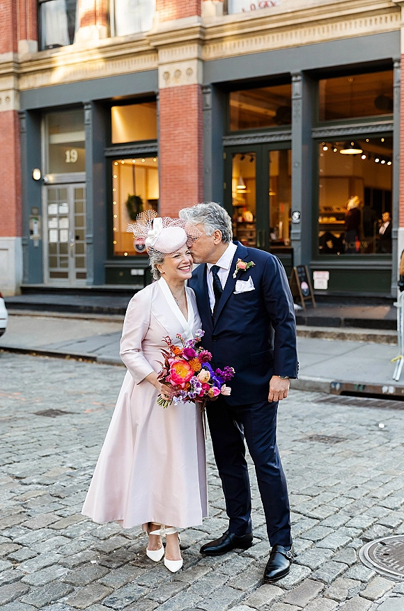 Wedding couple portrait of groom kissing bride’s forehead, her birdcage veil and bouquet against a cobblestone city street and brick facade