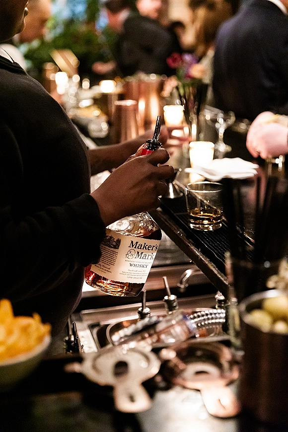 Wedding bar service with whiskey bottle and rocks glass on the counter, votive candles glowing as reception guests mingle in warm light