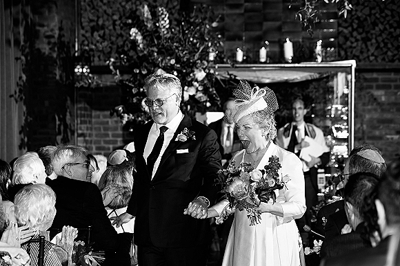 Wedding recessional as bride and groom walk hand in hand down the aisle, bride holding bouquet and birdcage veil, guests by brick fireplace