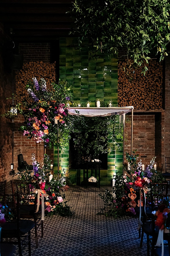 Ceremony backdrop with an indoor ceremony backdrop around a green tile fireplace, colorful florals, greenery garlands, candles and lanterns on brick floor