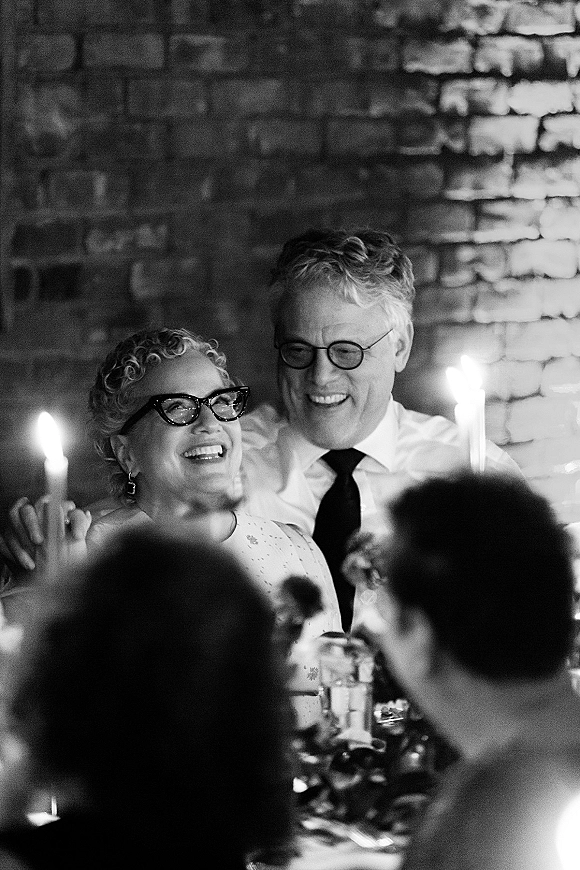 Wedding reception toast as the couple laughs during a wedding speech moment, lit by taper candles at a banquet table with brick wall behind