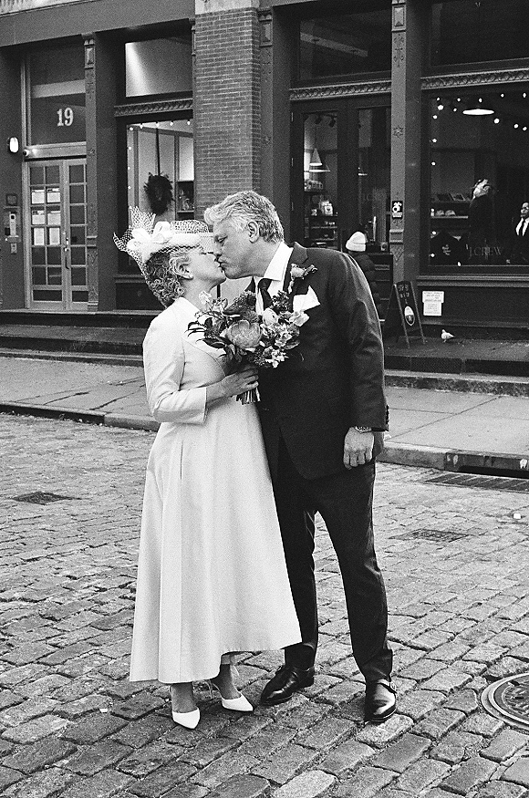 Wedding kiss portrait of a mature bride and groom kissing on a cobblestone city street, bride in birdcage veil holding bouquet by brick storefront windows