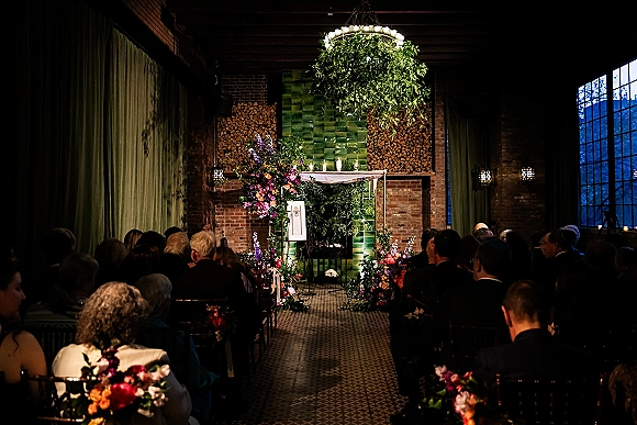 Indoor ceremony setup with an industrial wedding ceremony arch, lush florals and candles, against brick walls and large loft windows.