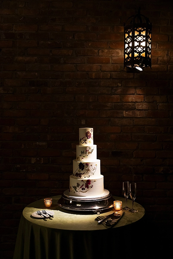 Wedding cake on a silver stand, four tier wedding cake with hand-painted florals, surrounded by votive candles and champagne flutes by a brick wall