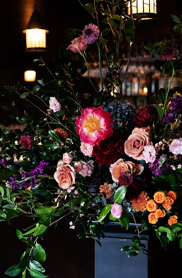 Wedding floral arrangement with roses and peonies in pink, burgundy, and orange on a black pedestal, set against warm lantern-lit bar shelves