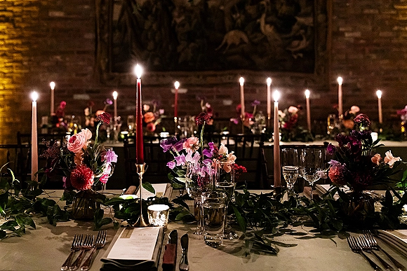 Reception tablescape with candlelit wedding reception taper candles in brass candlesticks, florals, greenery garland, and menus against a brick wall.