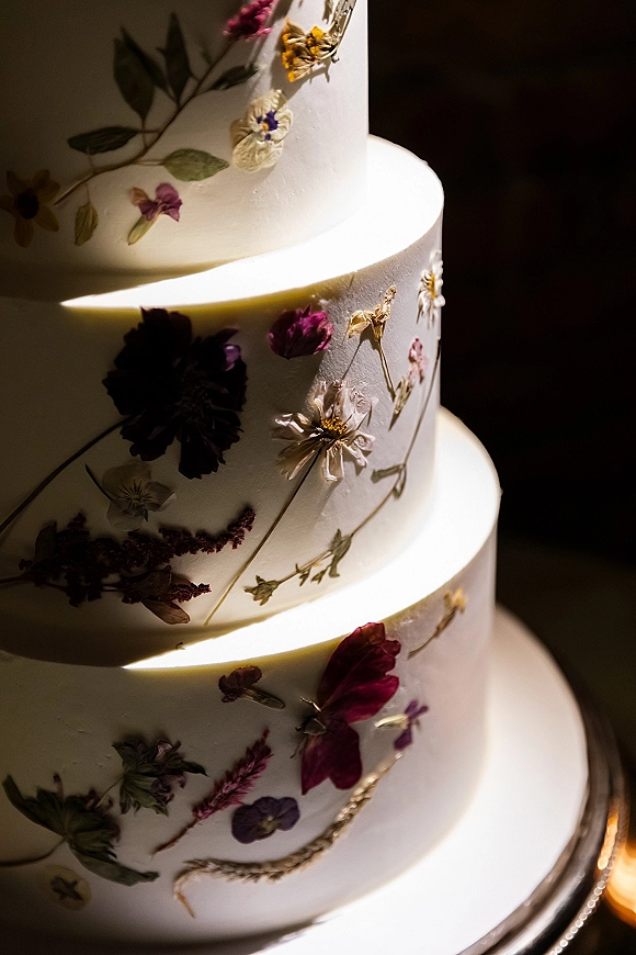 Wedding cake with pressed flower details on white fondant tiers, featuring floral appliques and botanical accents on a stand against dark backdrop