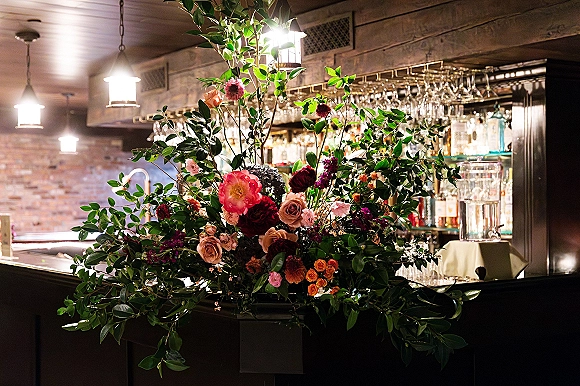 Wedding floral centerpiece with lush greenery and roses on a bar, set against brick wall shelves under warm hanging lantern lights