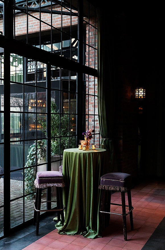 Cocktail table decor with green cocktail table linens, bud vase flowers, votive candles and place card by brick wall and industrial windows