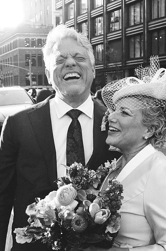 Wedding couple portrait in a black and white wedding photo, laughing together on a city street, bride in fascinator with birdcage veil holding bouquet