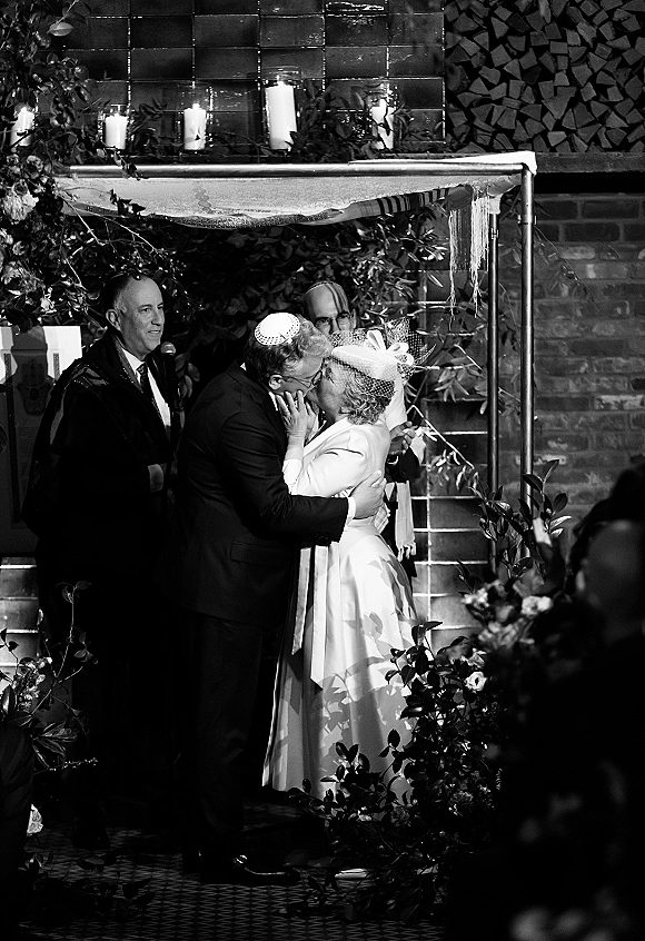 Wedding kiss under a chuppah as the couple embraces, bride in birdcage veil and fascinator, groom in kippah, candlelit brick-wall venue