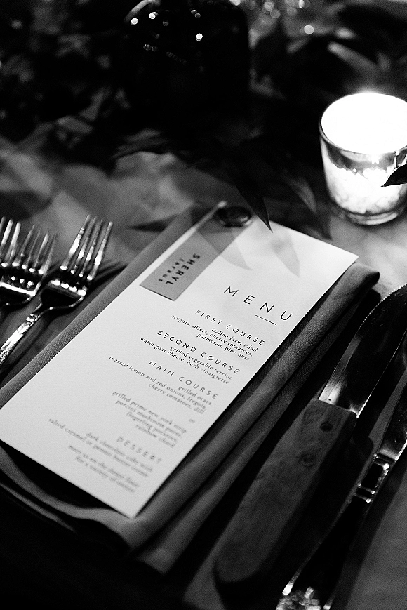 Wedding menu card with reception menu stationery tied by ribbon on a napkin, beside forks and knife, with votive candle and greenery on a low-light reception table