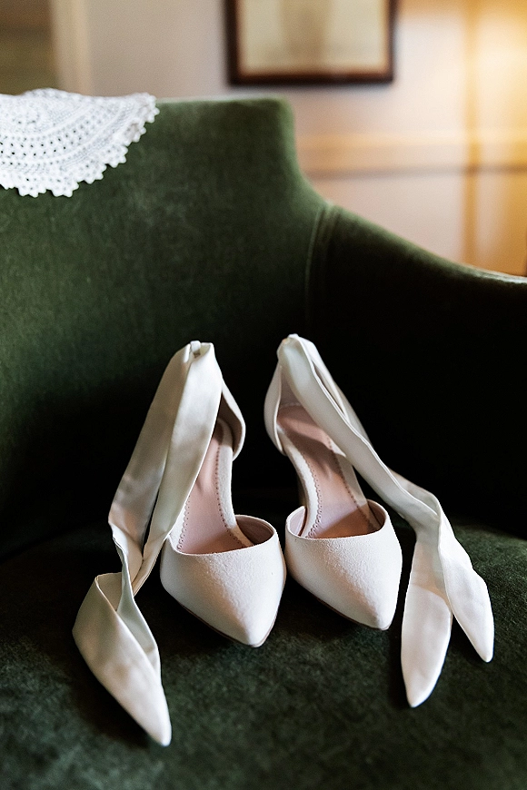 Bridal shoes, white wedding heels with pointed toes and ankle bows resting on a lace doily atop a green velvet chair indoors