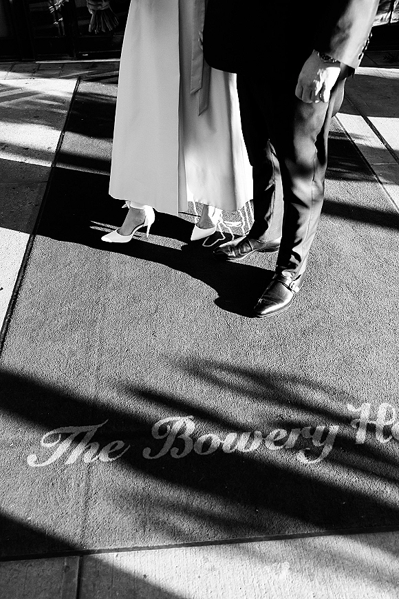 Couple portrait in a black and white wedding photo focusing on bride’s heels and dress hem beside groom’s shoes at a doorway doormat