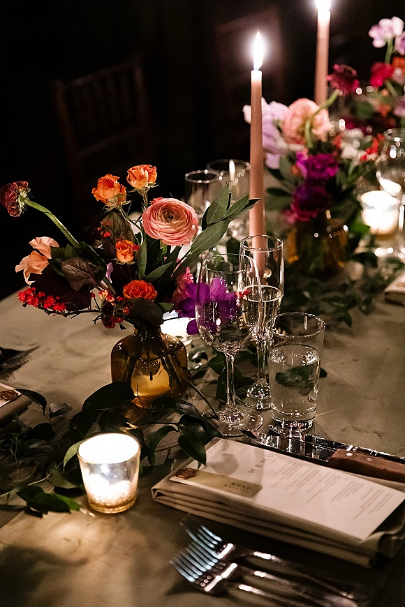 Reception tablescape with wedding table centerpiece of taper candles and lush florals in an amber glass vase, set in a moody dark room