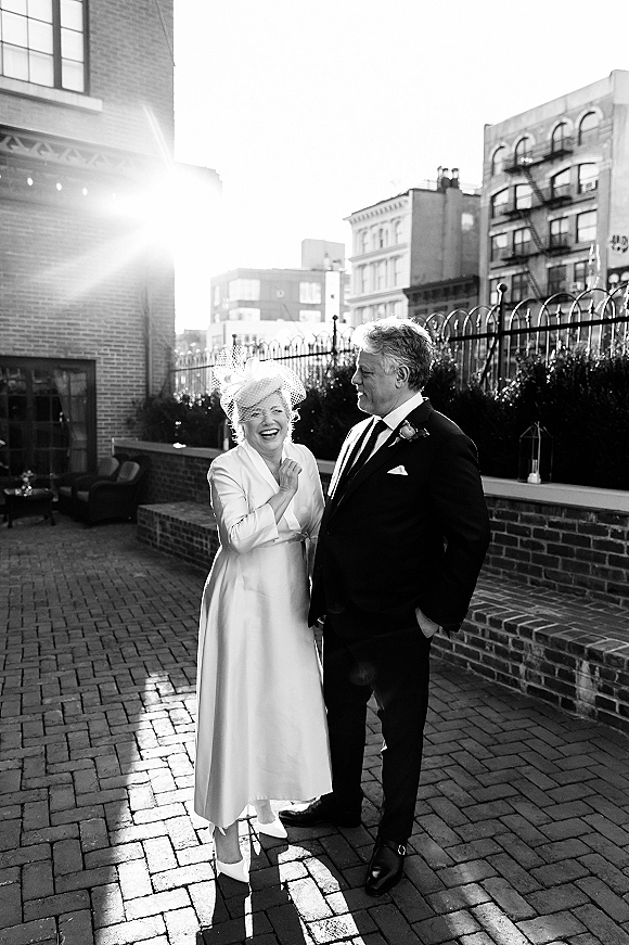 Couple portrait in a black and white wedding portrait style, bride in birdcage veil laughing as groom looks at her on a city rooftop terrace