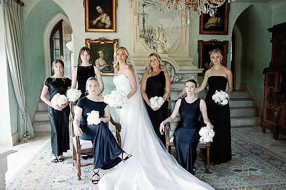 Bridal party portrait with bride and bridesmaids in black dresses holding white bouquets, posed by an ornate fireplace in a vintage room