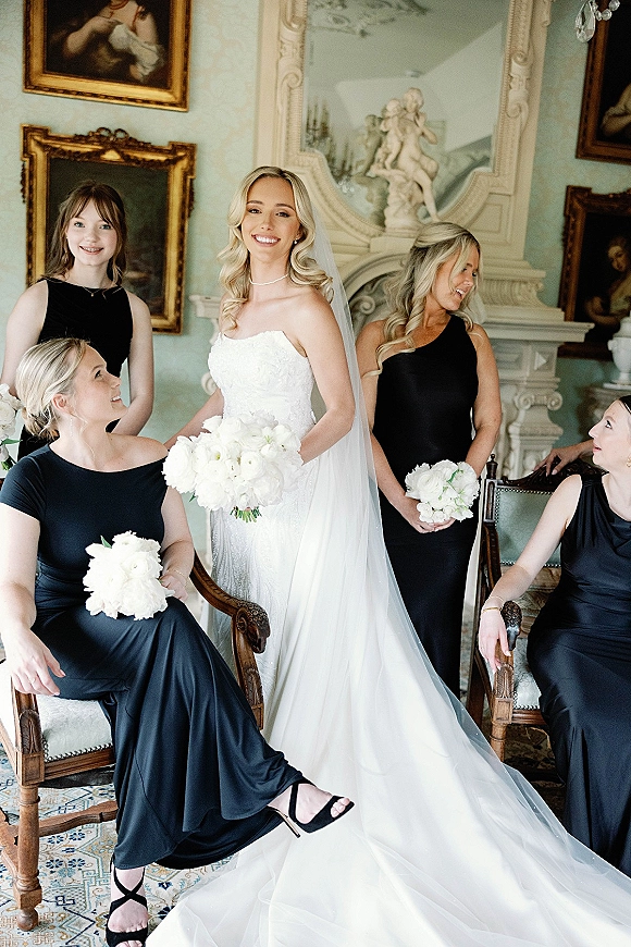Bride with bridesmaids in black dresses holding white rose bouquets, posed in a vintage room with ornate fireplace and chandelier backdrop