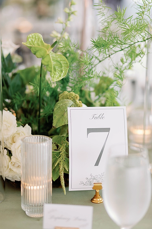 Wedding tablescape with wedding table number card in a gold holder, lush greenery and white flowers, ribbed glass candle, blurred reception background