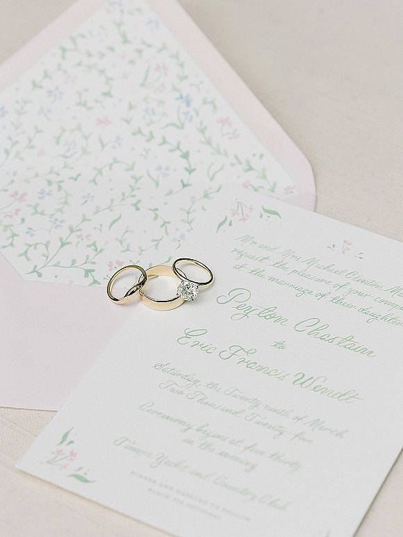 Wedding rings with diamond engagement ring and bands resting on a calligraphy wedding invitation atop soft white fabric flat lay