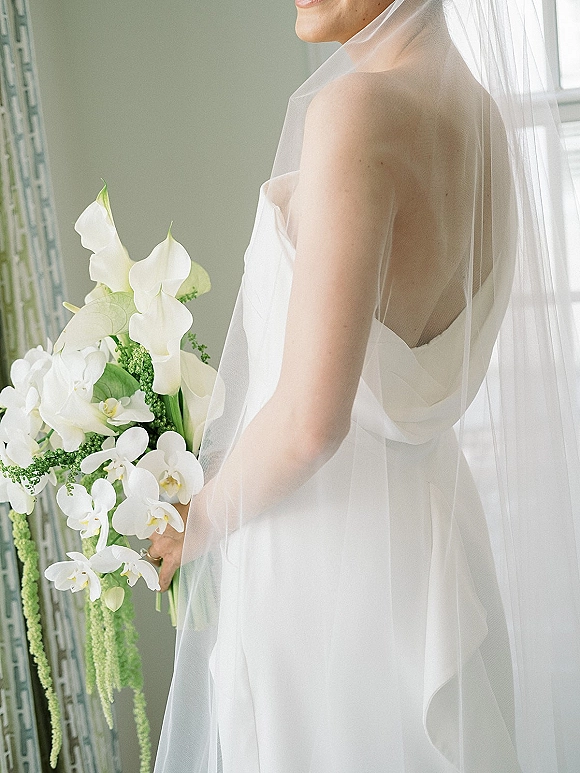Bridal portrait of a bride holding bouquet of white calla lilies and orchids, veil over shoulders in soft window light indoors
