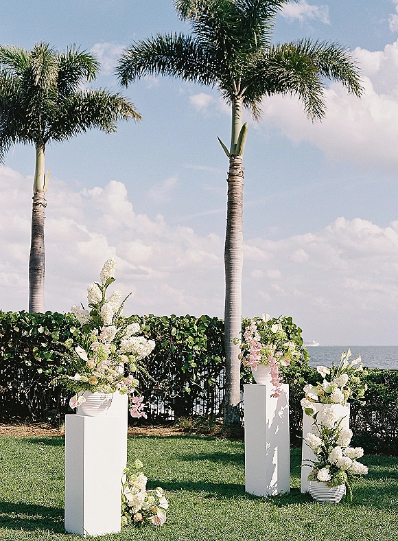 Ceremony floral pedestals and wedding floral pillars with white and green arrangements on a lawn, set against palm trees and ocean sky