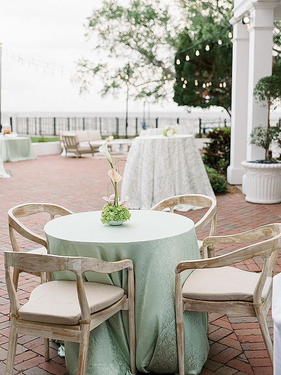 Reception tablescape with an outdoor wedding reception table dressed in sage green linen, calla lily and hydrangea centerpiece under string lights by a waterfront patio